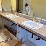 Old double sink vanity with outdated tile countertop and gold fixtures during bathroom demolition.